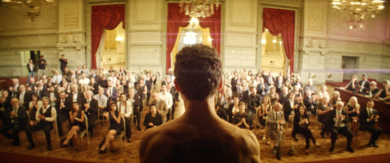 A naked man stands infront of an audience in a concert hall