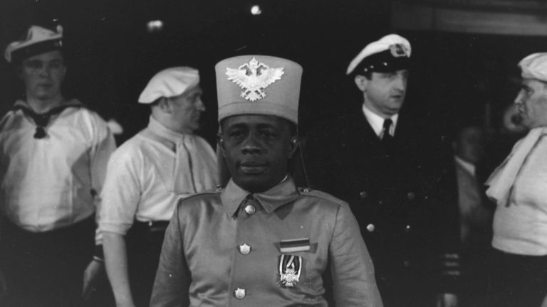 Filmstill "Majubs Reise", ein Mann steht in einer Uniform vor einer Reihe anderer Männer in Uniform