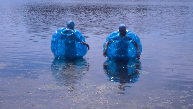 Zwei Menschen stehen im Wasser. Sie sind in blaue Plastikkugeln gehüllt