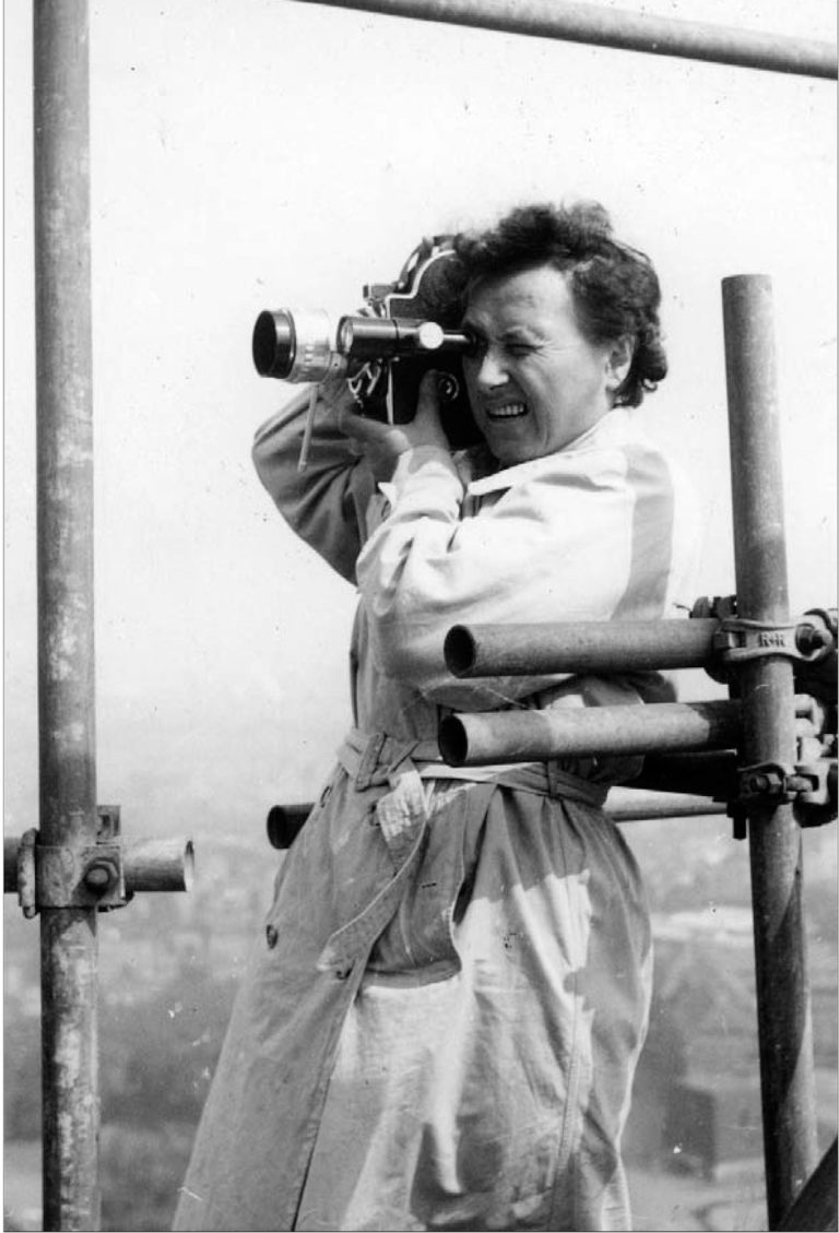 Elisabeth Wilms steht im Trenchcoat und mit der Filmkamera auf der Schulter auf einem Baugerüst