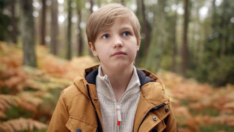 The picture shows a boy in a brown anorak in close-up. He is standing in the forest