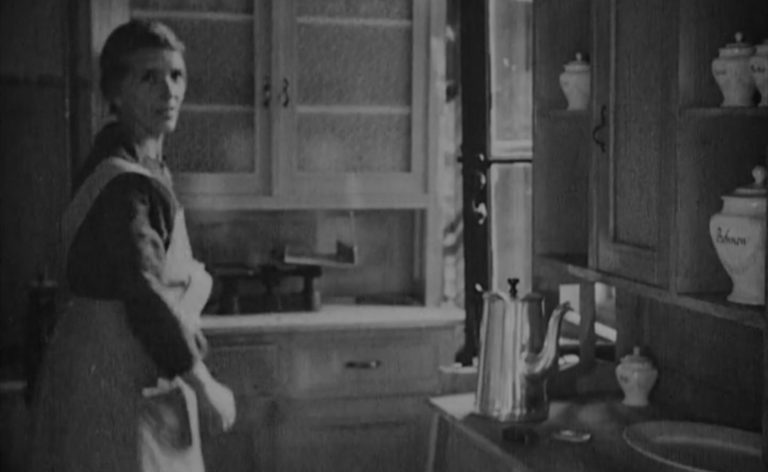 Schwarz-weiß-Aufnahme einer Frau die in einer Küche steht und in Kamerarichtung blickt.