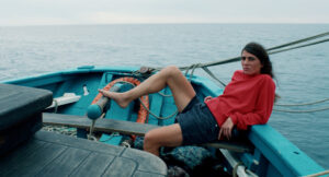 Das Filmstill zeigt eine Frau, die sich an den Rand eines Bootes öehnt und mit dem Fuß das Boot steuert.