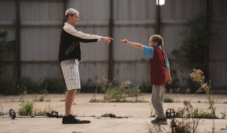 Filmstill aus dem Film Georgie. Ein jugendlicher Junge und ein junges Mädchen stehen sich vor einer Halle gegenüber und berühren mit ausgestreckten Zeigefinger fast ihre Hände.