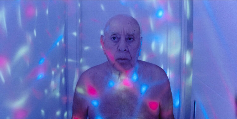 Ein älterer Mann schaut ernst an der Kamera vorbei. Discoähnliche Lichter sind zu sehen und der ganze Raum erscheint in lilanem Licht.
