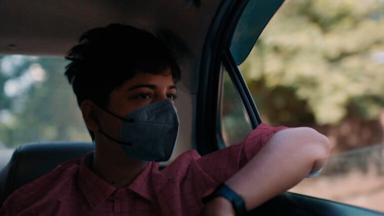 A young person wearing an FFP2 mask is sitting in the back seat of a car with one arm leaning out of the open window and looking out.