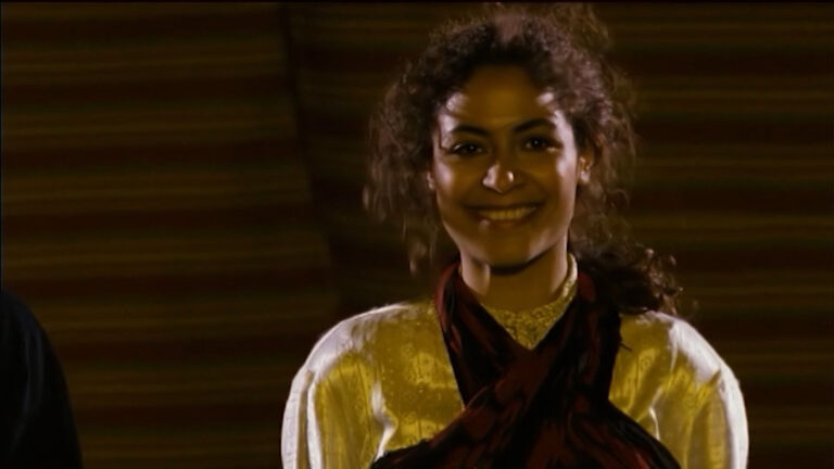 A woman with brown curls in a golden top looks into the camera with a smile.