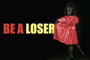 Eine Person steht in einem roten Lackmantel mit dem Rücken zur Kamera. Auf dem Kopf trägt sie eine Affenmaske und blickt in die Kamera. Neben der Person steht der Text "Be a Loser".