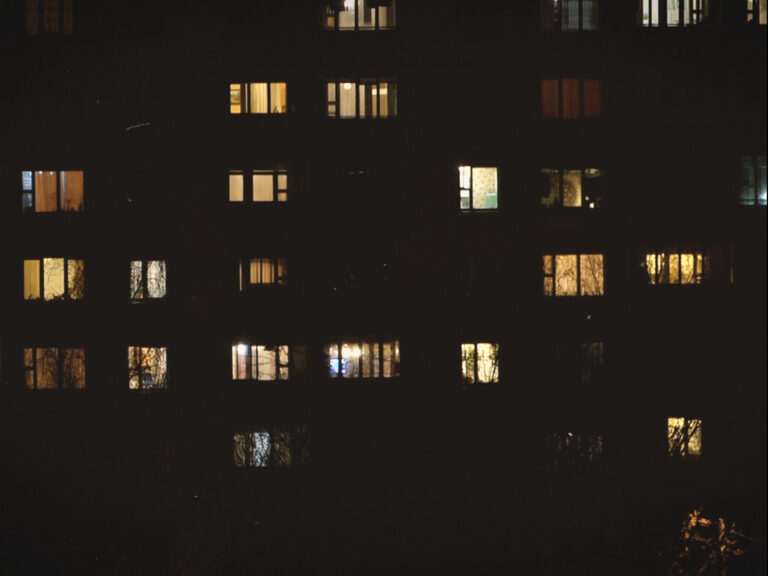 Zu sehen ist eine Häuserfront bei Nacht mit hell erleuchteten Fenstern.