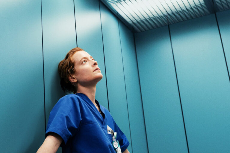 A woman in surgical scrubs stands pensively leaning against the wall in an elevator.