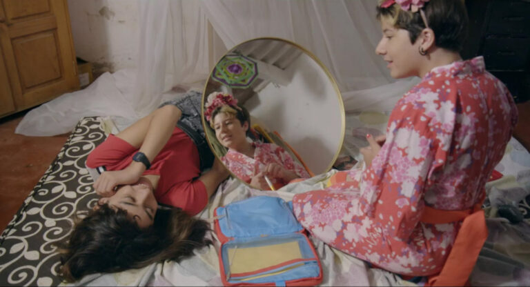 2 Personen liegen und sitzen nebeneinander auf dem Bett. Vor der einen Person steht ein Spiegel und ein Rucksack liegt zwischen den beiden Personen auf dem Bett.