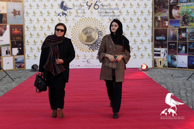 Zwei Frauen mit dunkler Kleidung und Kopftuch laufen über einen roten Teppich, im Hintergrund steht eine weiße Fotowand.