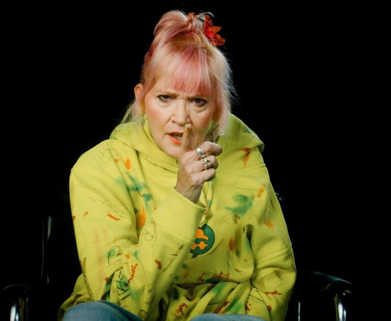 A woman with pink hair and a green jacket is looking towards the camera with her hand raised.