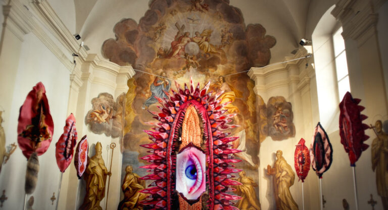 Heller Innenraum einer Kirche, an einem Seil hängt eine große gebastelte pinke und rosane Vulva in deren Mitte in großes Auge geklebt ist.