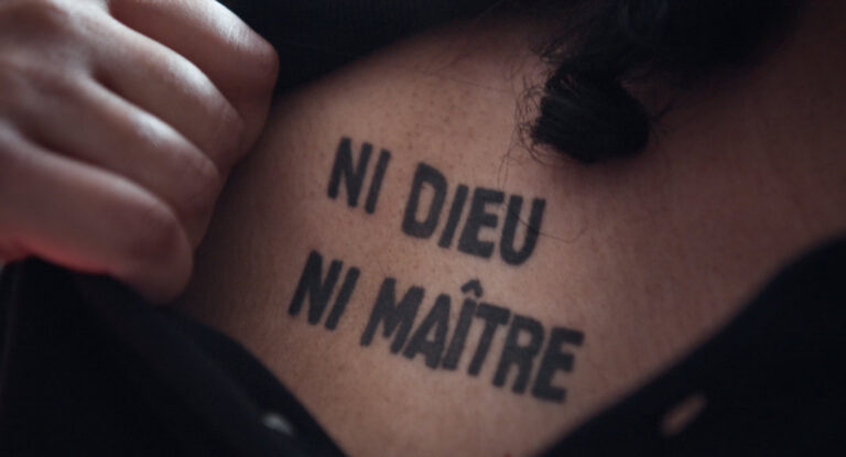 Eine Hand zieht ein Stück Stoff zur Seite, sodass ein Nackentatoo mit den Worten "Ni Dieu Ni Maitre" sichtbar wird.